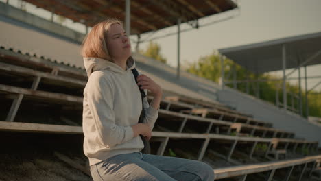 back view of a woman in casual attire holding black bag as she walks along rusty bleachers in an old stadium and sits down as the sun reflect off her