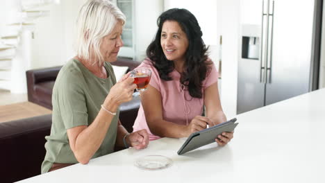 Felices-Y-Diversas-Mujeres-Mayores-Usando-Tabletas-Y-Bebiendo-Té-En-Una-Cocina-Soleada,-Cámara-Lenta