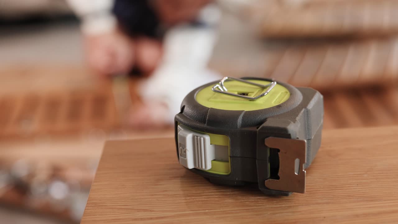 A tape measure rests on a wooden surface with a blurred background, suggesting a furniture assembly setting