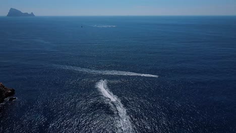 dolly forward aerial shot of two jet skis enjoying the crystal clear waters of sa fig, on the island of ibiza in spain with islands on the horizon in the mediterranean sea