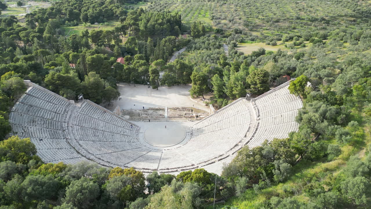 tomada aérea del teatro de epidauro en grecia, rodeado de exuberante vegetación con una sola persona de pie en él, destacando la inmensidad de la antigua estructura