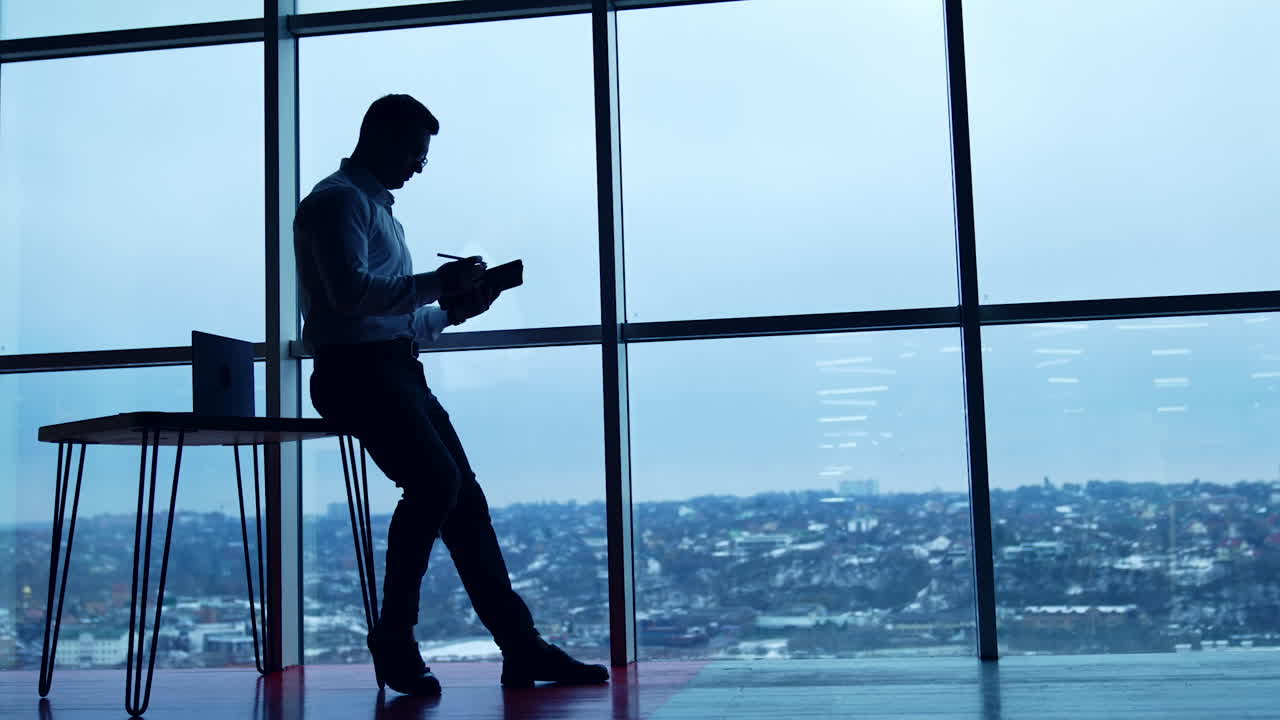 Businessman sitting on desk draws with a pen on his I-pad. Silhouette of an employee working in office with panoramic window. Low angle view.