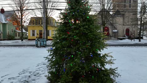 Slow-aerial-rising-shot-of-decorated-Christmas-tree-revealing-church,-houses,-and-beautiful-golden-hour-sunburst-light-on-snow-covered-day-in-Wellsboro-PA