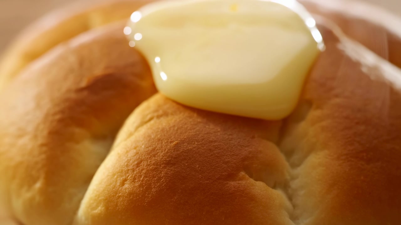 Vue macro extrême d'un petit pain tressé doré avec un nappage jaune brillant s'étalant sur la crête centrale, formant des flaques réfléchissantes, des reflets lumineux, une texture de boulangerie chaleureuse et un détail appétissant