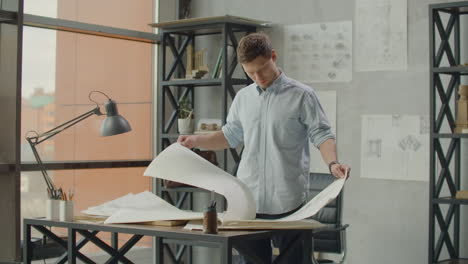 bearded man hipster standing faces to panoramic window and looks at blueprints. young architect in stylish loft office works with documents. creative architect thinking on the big drawings.