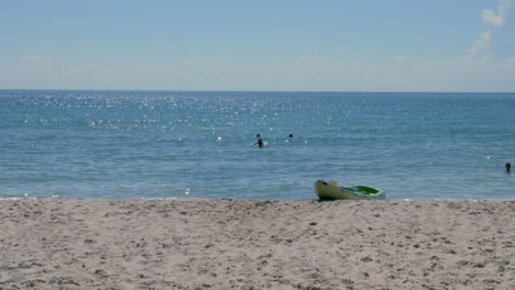An-empty-kayak-sits-on-the-shore-of-a-beach-as-people-swim-in-the-distance