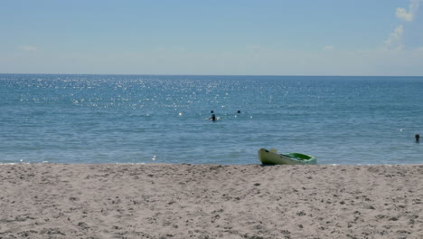 an empty kayak sits on the shore of a beach as people swim in the distance