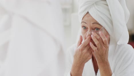 happy senior caucasian woman looking at mirror in bathroom and applying cream on her face