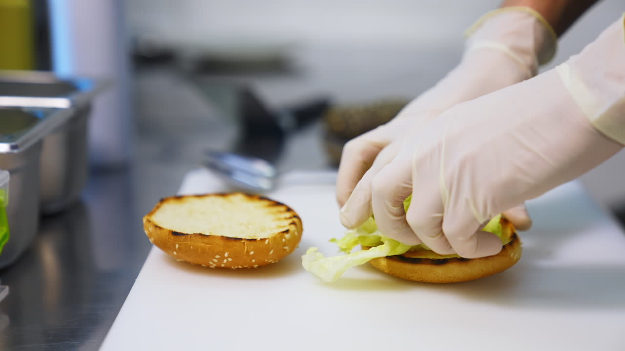 Hands in latex gloves put lettuce on top of fried bun. Making burger on white table. Fast food concept.