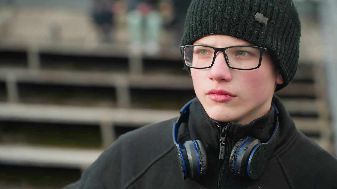 Close-up of a young boy wearing a black outfit and beanie, looking focused and contemplative, showing his glasses and a pair of headphones resting around his neck