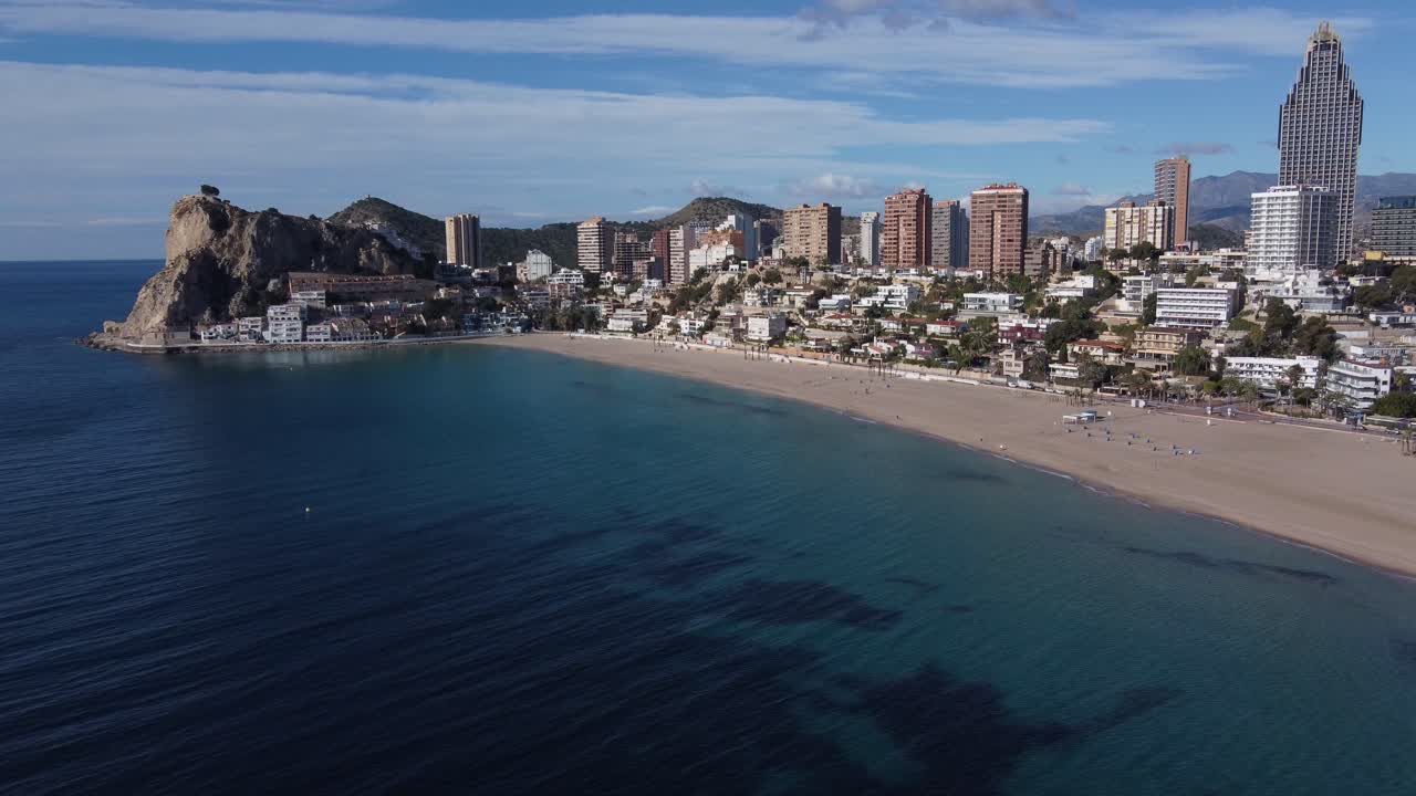 Drone shot of crystal clear blue water, sandy beach and luxury buildings on hill in Benidorm City, Alicante