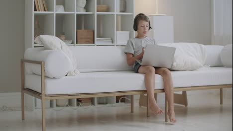 Fröhlicher-Junge-Kommuniziert-Mit-Jemandem-Per-Videoanruf-Auf-Dem-Laptop-über-Kopfhörer,-Der-Zu-Hause-Im-Bett-Sitzt