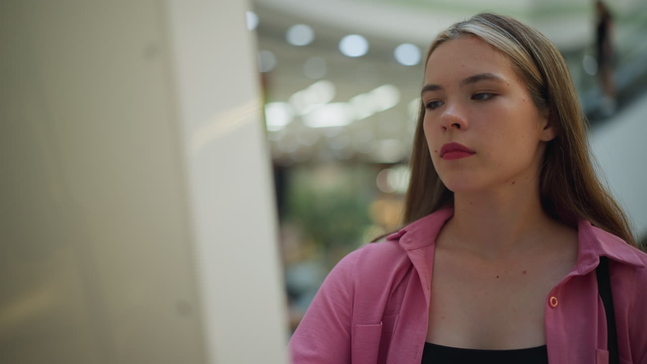 dama en vestido rosa con bolso negro intensamente enfocado en el uso de una pantalla en un centro comercial bien iluminado, el fondo cuenta con luces borrosas y escaleras mecánicas con personas en él, mientras que ella parece centrarse
