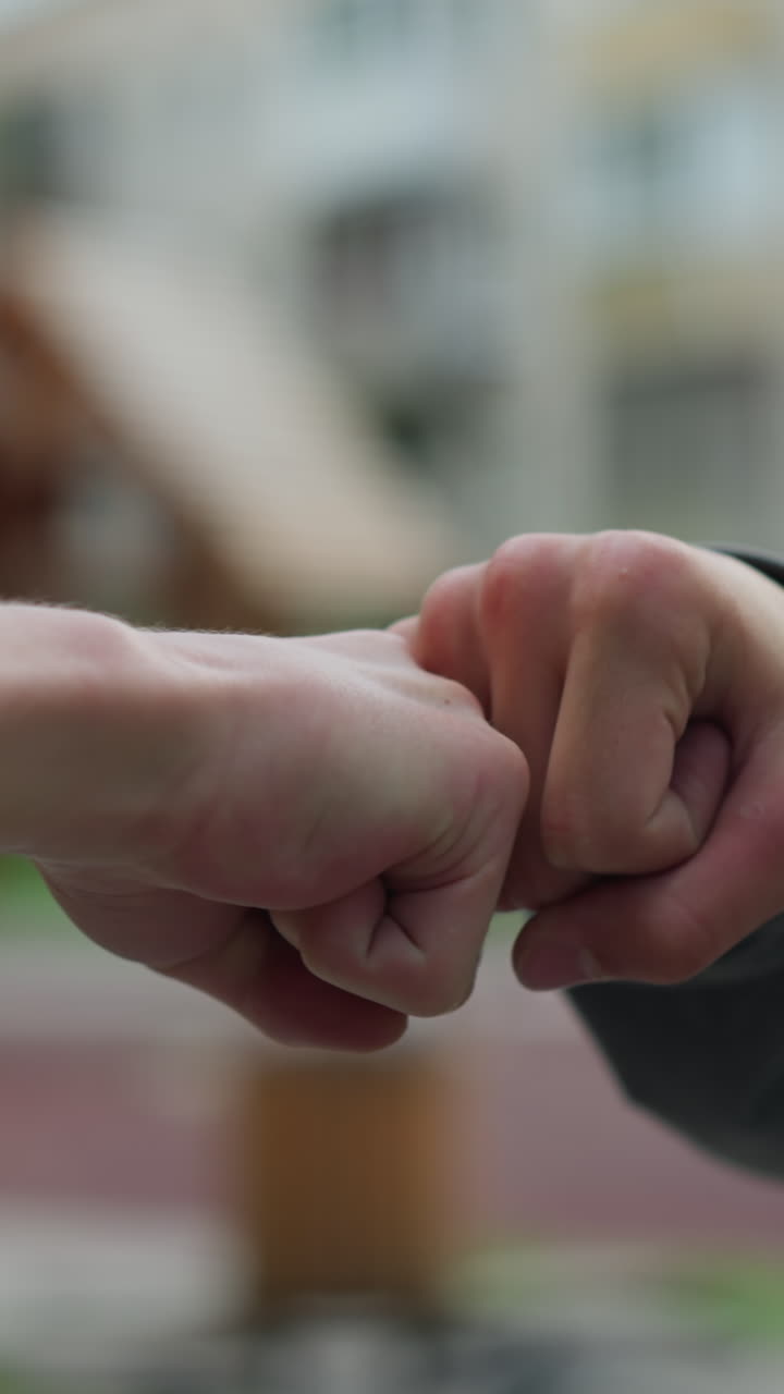 primer plano de manos que interactúan en un golpe de nudillo lúdico, una persona con una camiseta gris extiende su mano para conectarse con otra, con un fondo borroso con edificios residenciales