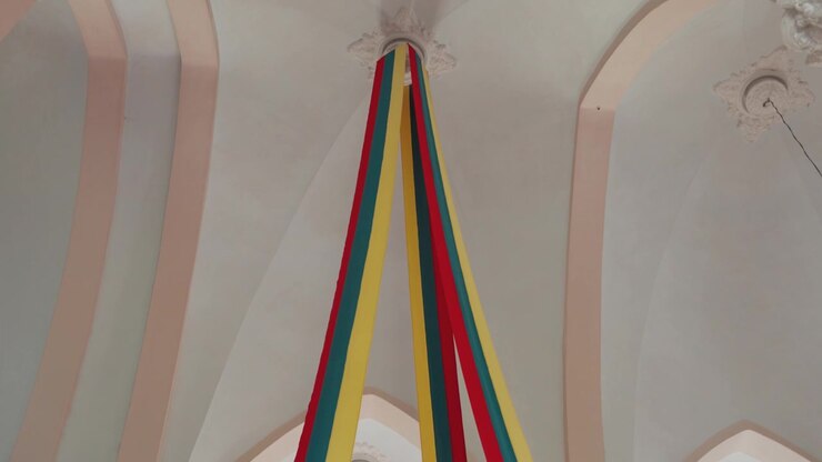 Vivid red, yellow, and blue ribbons cascade from a high vaulted ceiling in a softly lit, ornate historic building. The smooth camera tilt highlights elegant columns and intricate moldings.