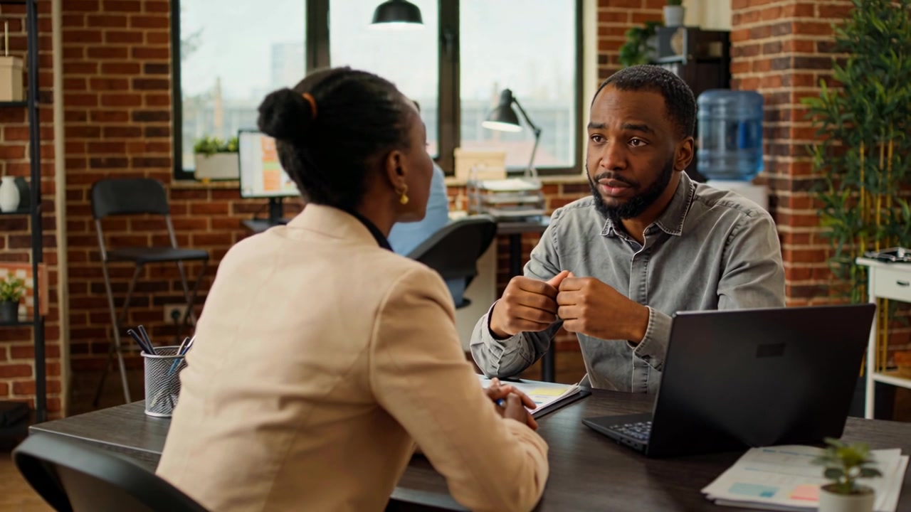Starting gray-shirted man leaning and explaining at loft desk with laptop papers, seeking agreement