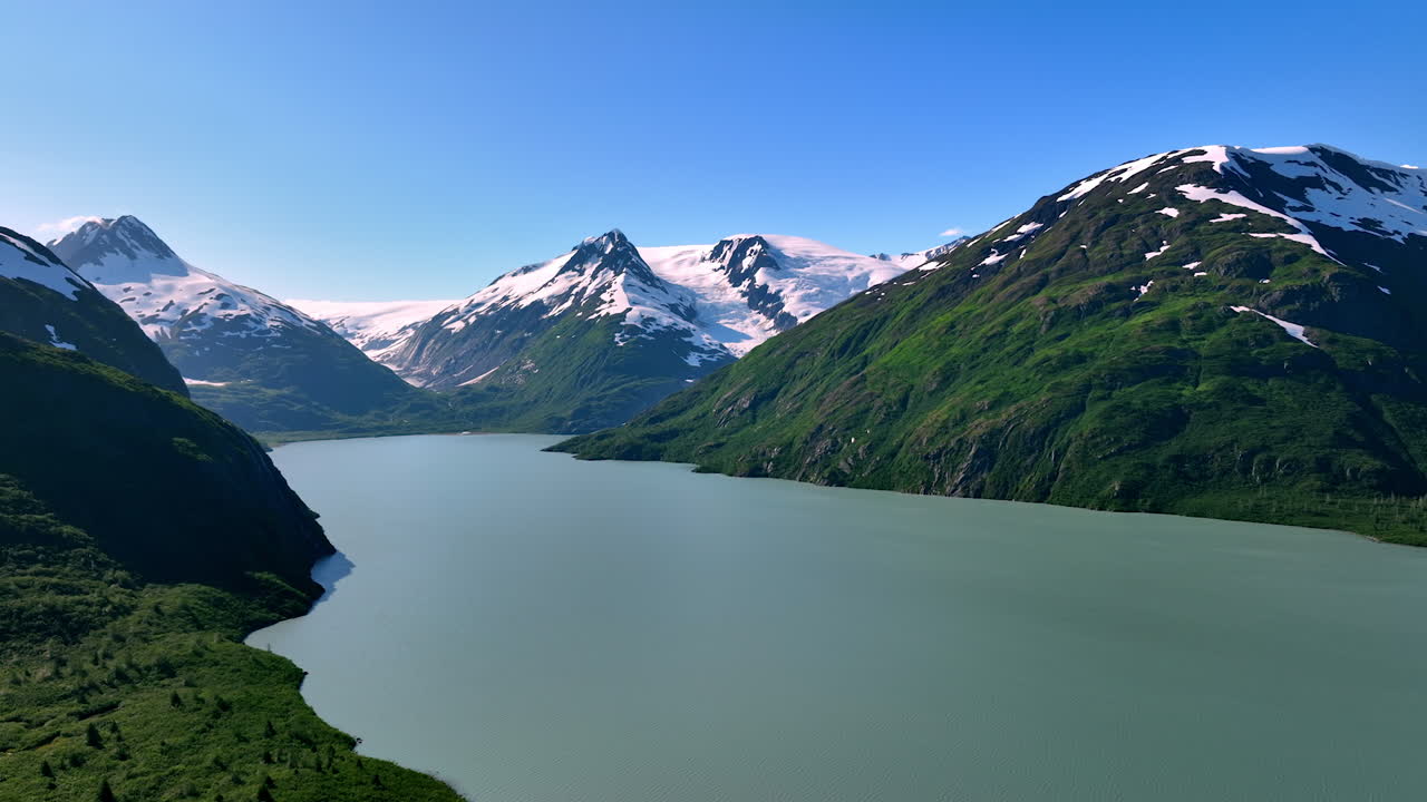 Alaska serene waters shine. Majestic mountains with snow capped peaks surround a peaceful lake in Alaska under a clear blue sky, perfect for travel