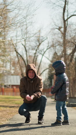 un padre, vestido con una chaqueta marrón, se agacha en un sendero del parque mientras libera algunas partículas en el aire, mientras que su hijo pequeño, con una brillante chaqueta negra y vaqueros, corre emocionado para atraparlos