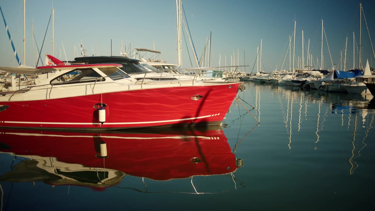 Motoscafo rosso ormeggiato in un porto turistico affollato, il suo colore vivace che si riflette sull'acqua calma sotto un cielo azzurro limpido.