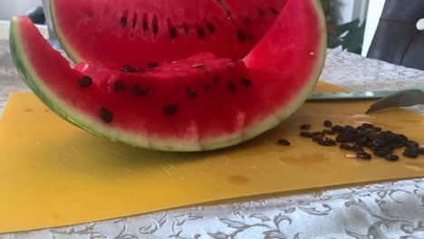 Nahaufnahme-Einer-Geschnittenen-Wassermelone-Mit-Leuchtend-Rotem-Fruchtfleisch-Und-Schwarzen-Kernen-Auf-Einem-Gelben-Schneidebrett-Neben-Messer-Und-Gabel