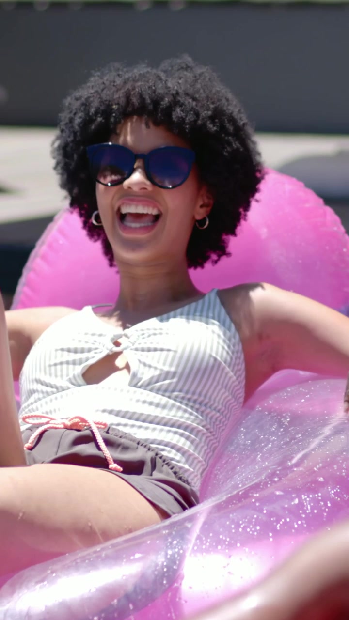 Vertical video: Smiling woman on pink float raising arms poolside in sun, friends wearing swimwear