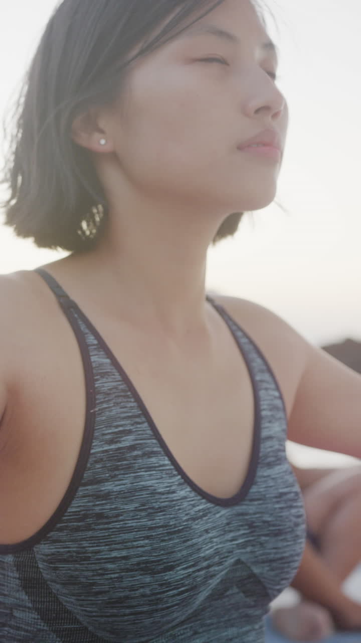 Vertical video of happy biracial couple doing yoga and meditating at beach, in slow motion