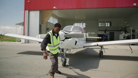 african american technician towing light aircraft from hangar at airfield