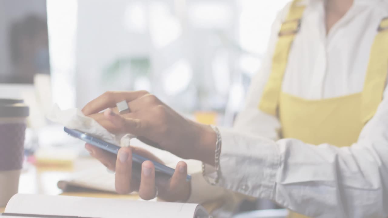 Animation of flashing words with woman disinfecting smartphone in office wearing face masks