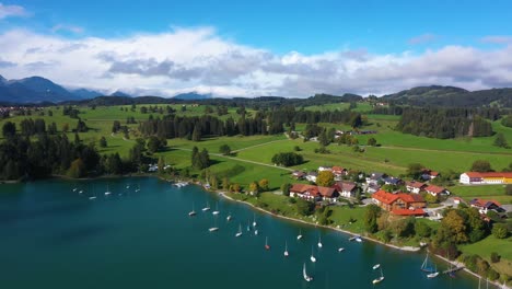 Increíble-Vista-Aérea-Que-Desciende-Cerca-De-Un-Enorme-Lago-Con-Vistas-A-Un-Pequeño-Pueblo-Con-Barcos-En-Forggensee-En-Baviera,-Alemania-En-4k