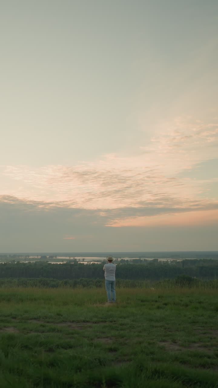 un hombre adulto, con una camisa blanca, sombrero y vaqueros, se encuentra contemplativo en un campo cubierto de hierba junto a un lago tranquilo al atardecer. el paisaje sereno y la luz cálida crean una atmósfera pacífica y reflexiva