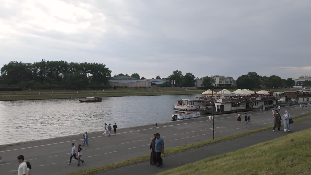 Scenic view of people strolling along the River Vistula promenade in Krakow, Poland, with boats and lush greenery under a cloudy sky. Pan right