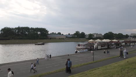 scenic view of people strolling along the river vistula promenade in krakow, poland, with boats and lush greenery under a cloudy sky. pan right
