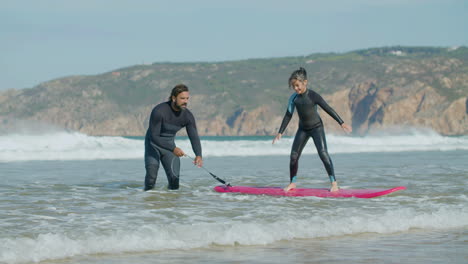 Lange-Aufnahme-Eines-Glücklichen-Mädchens,-Das-Mit-Hilfe-Eines-Trainers-Auf-Einem-Surfbrett-Steht