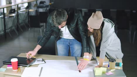 Young-man-and-woman-working-on-a-project,-drawing-and-thinking-in-modern-office.-Using-compass.-Coffee-cup-and-laptop-on-the