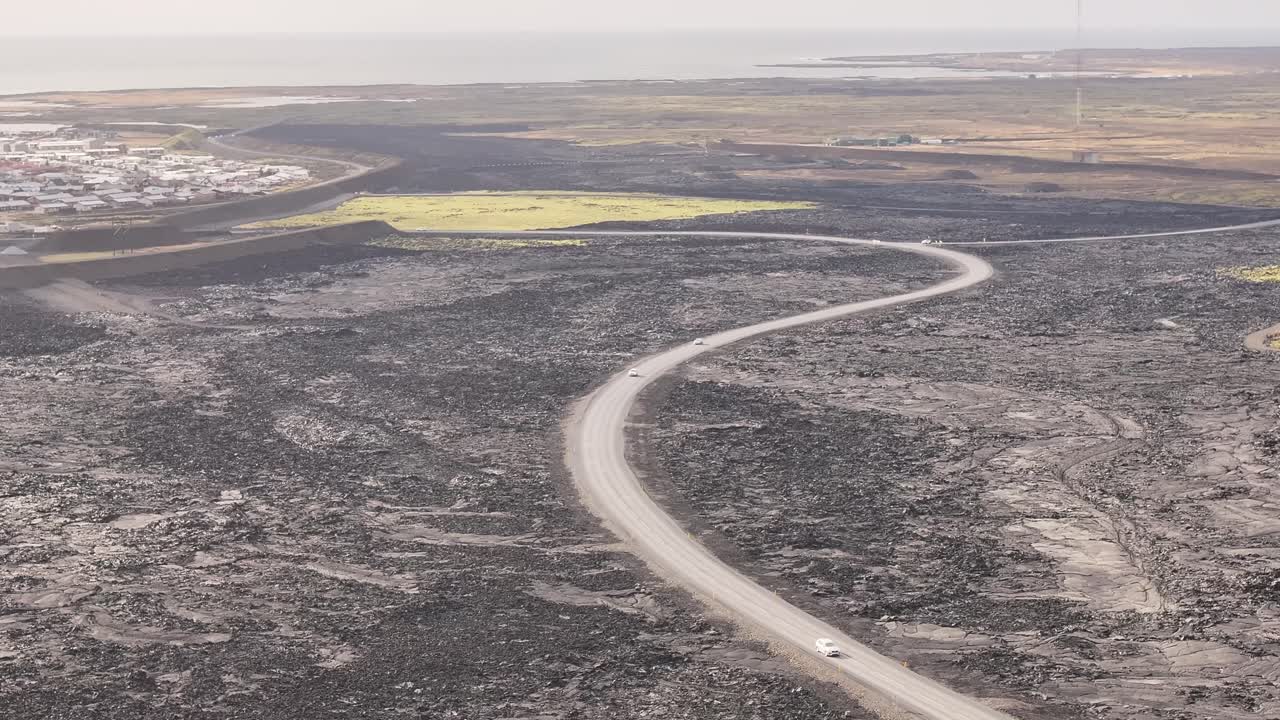 A barren, desolate Icelandic landscape featuring a winding road cutting through vast, rugged terrain, dark volcanic ground, sparse vegetation, and distant mountains under an expansive sky.