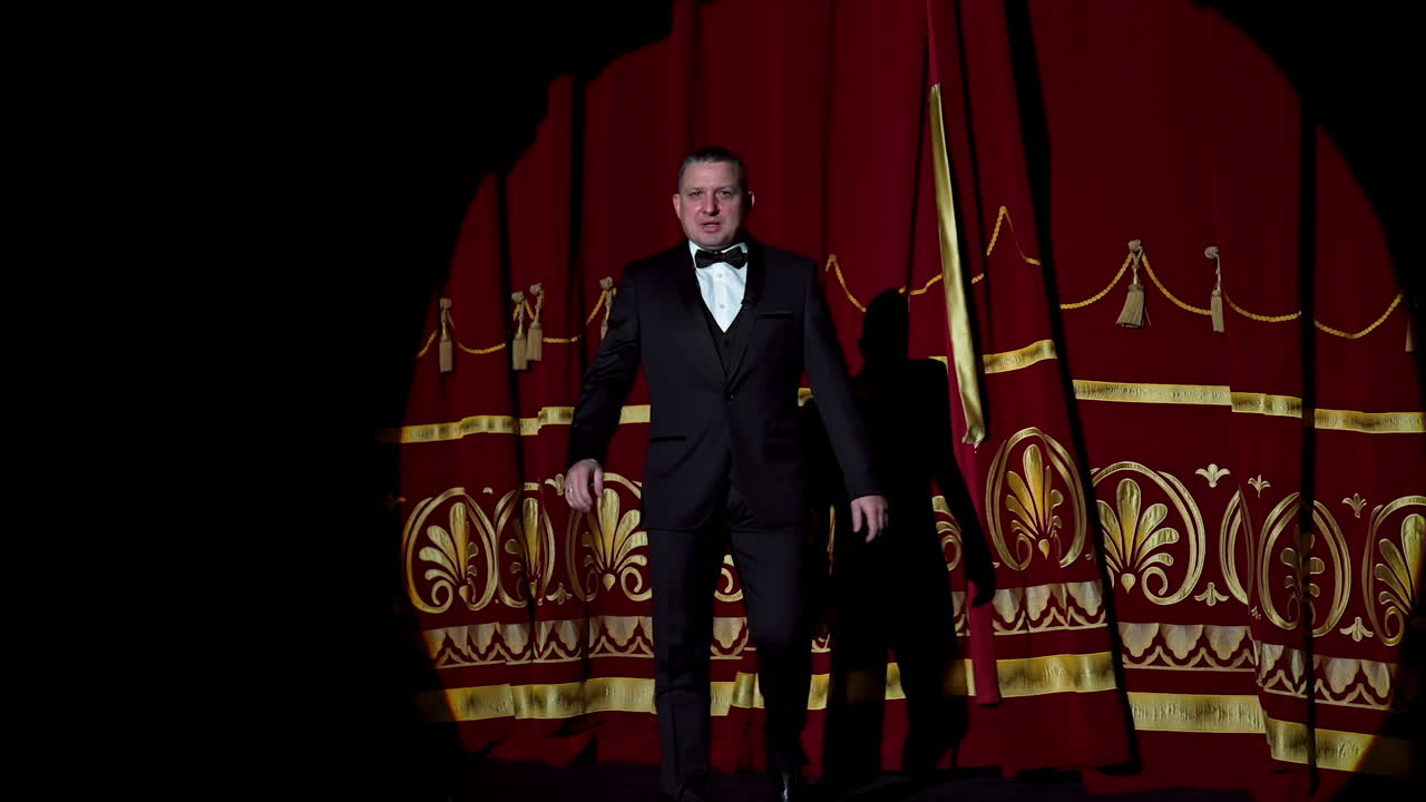 Man is speaking in the light of the floodlights standing on stage to the camera. Actor, artist on performance. Active gestures with hands.