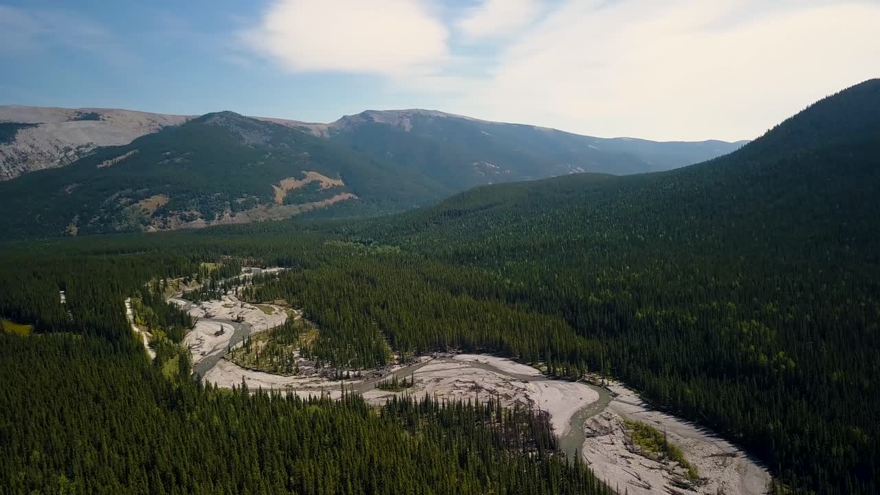 Aerial Daytime Wide Shot Moving Right Fast And Turning Left To Reveal Bends Of A Swift Steep River Flowing Through A Summer Pine Forest Down A Valley Between Rocky Mountain Peaks in Alberta Canada