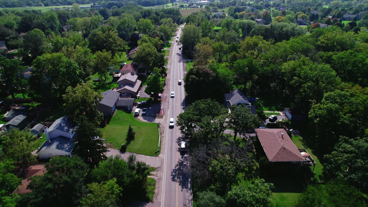 Aerial drone shot follows a straight road surrounded by American houses adorned with lush green gardens in an American neighborhood
