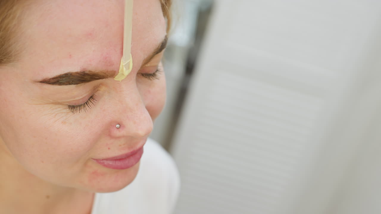 Close up of beautician applying wax to eyebrow of smiling client with closed eyes. Client appears relaxed and content while wearing nose stud during eyebrow grooming in bright salon environment