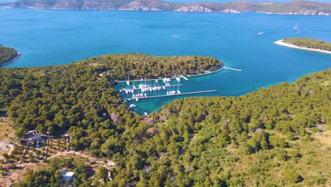 Yachts-dock-in-a-bay-near-the-coastal-city-of-Croatia-against-a-backdrop-of-blue-skies-and-blue-clear-water,-green-lush-trees-and-houses-with-red-roofs