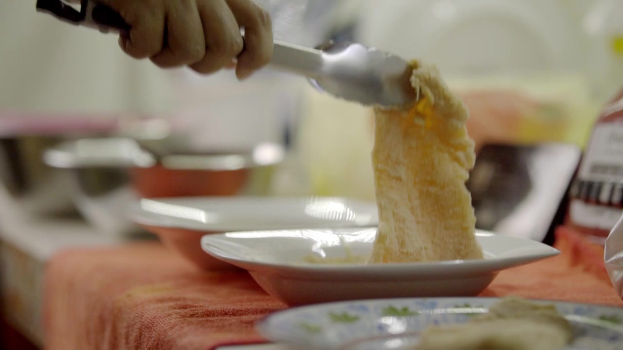Close-up of a hand using a fork to place cooked chicken onto a white plate. The serving action takes place at a dining table during meal preparation with dishes in the background.