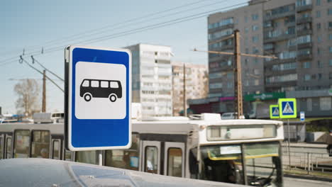 close view of a bus stop sign in an urban setting with buses and buildings in the background, with cars and a bus in motion