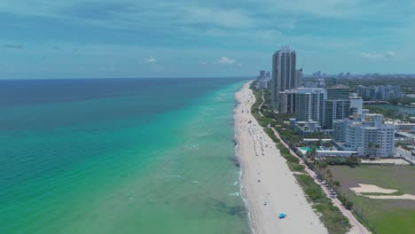 aerial flight over sandy beach with turquoise water of miami beach. luxury residential towers and houses in first row. people and tourist relaxing at beach. wide shot. florida, united stats