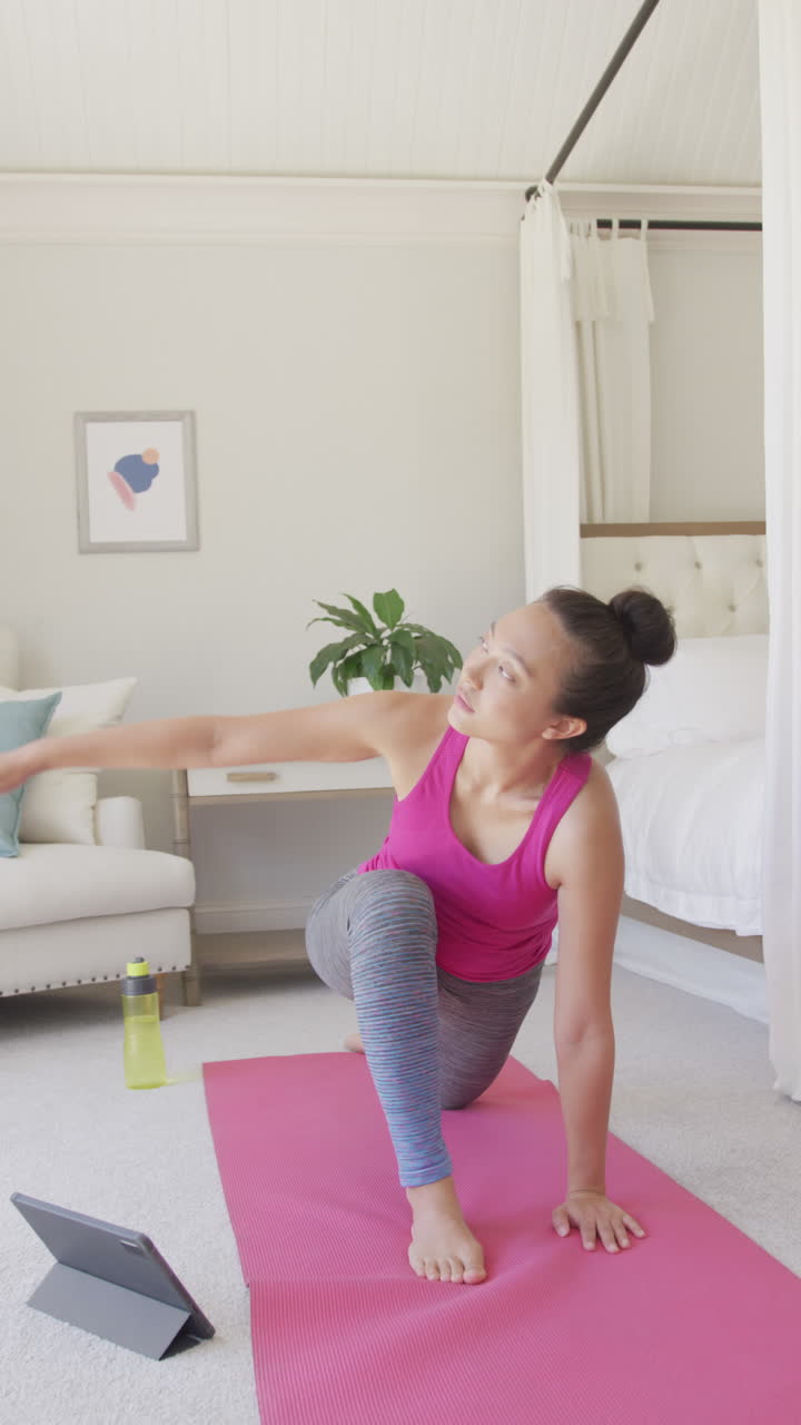 Vertical video of happy asian woman doing yoga and stretching in bedroom, in slow motion