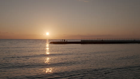 Lapso-De-Tiempo-De-La-Puesta-De-Sol-Sobre-El-Mar-Con-Cielo-Naranja-Nuboso-Y-Silueta-De-Personas-En-El-Muelle-Y-Barcos-En-El-Horizonte