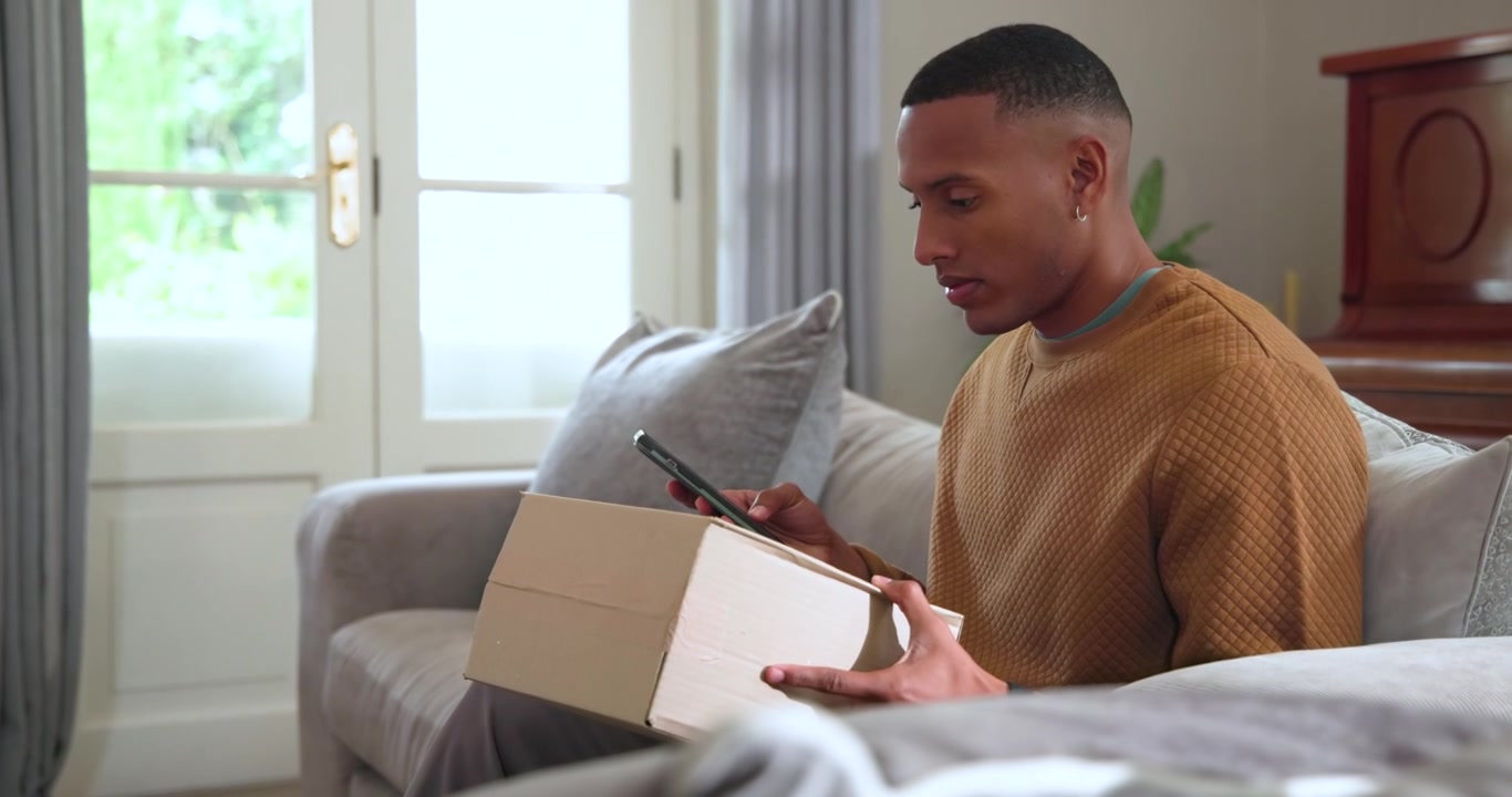 African American man after delivery alert sitting on gray couch opening box and checking smartphone