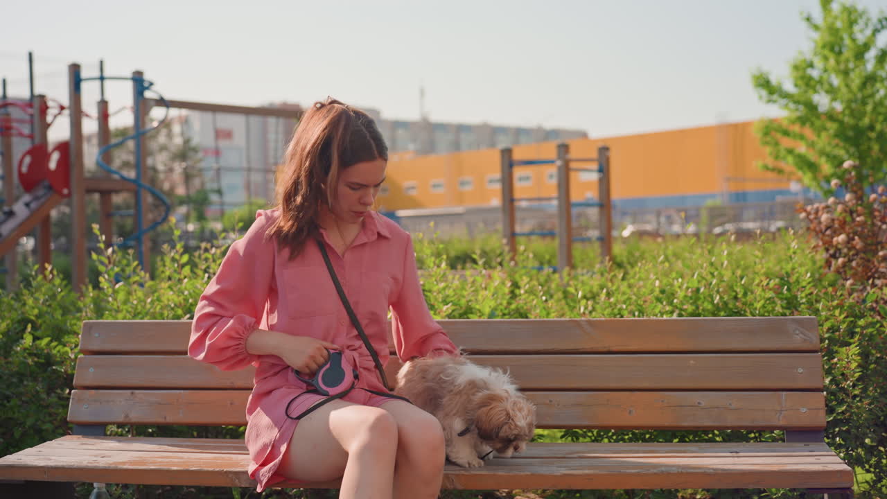 Young Caucasian Woman Petting Dog On Bench Near Playground Hedge, Gentle Grooming And Collar Check, Sunlit Wooden Seat, Calm Owner Moment, Relaxed Smile, Casual Outfit, Soft Fur Interaction, Urban