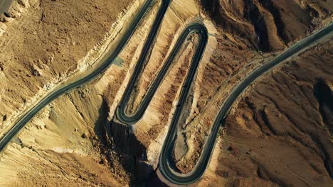 Four-vehicles-drive-downhill-on-the-black-asphalt-between-the-sharp-switchbacks-in-the-mountainous-landscape-of-sand-colored-Mitzpe-Ramon-in-Israel