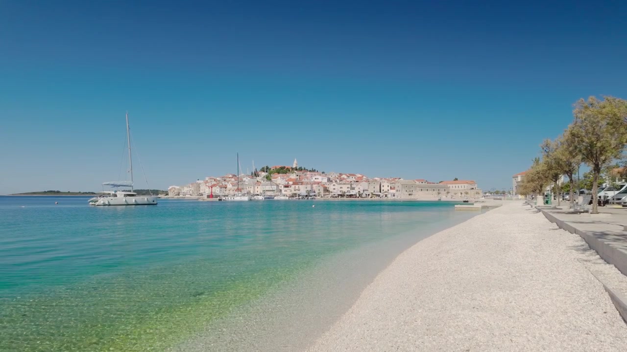 Ripresa a mano dalla spiaggia di Primošten che guarda verso la penisola del centro storico, con tranquille barche a vela sull'Adriatico luminoso e una dolce atmosfera estiva marittima