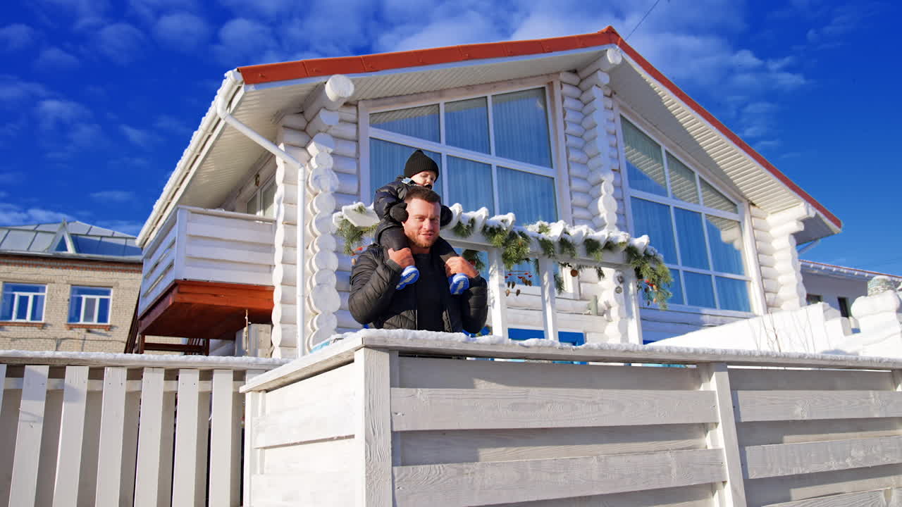 Smiling Caucasian man in black jacket holds a baby boy on his shoulders. Father and son near their house on sunny winter day. Low angle view.